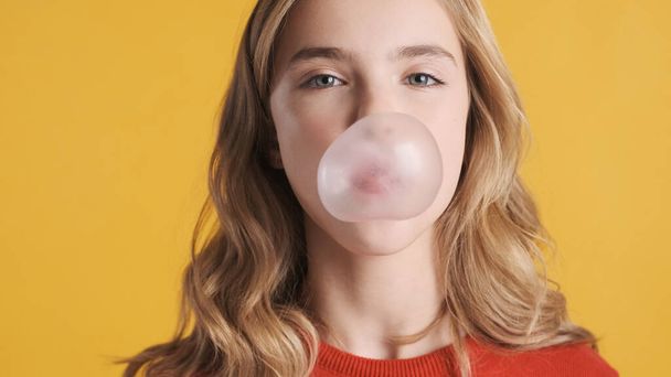 Close up pretty blond teenager dívka s vlnitými vlasy foukání bublina z žvýkačky na kameru přes žluté pozadí. Bonboniéra - Fotografie, Obrázek