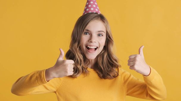 Docela pozitivní blond teenager dívka ve stranickém klobouku hledá šťastný vedení palce nahoru na kameru přes žluté pozadí - Fotografie, Obrázek