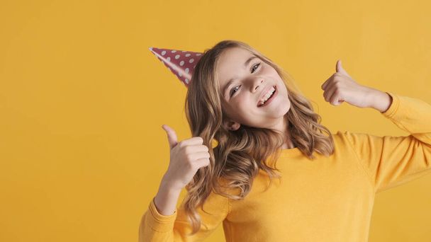 Positive blond teenage girl in party hat looking happy keeping thumbs up on camera isolated on yellow background. Cheerful girl like her birthday party - Foto, Imagen