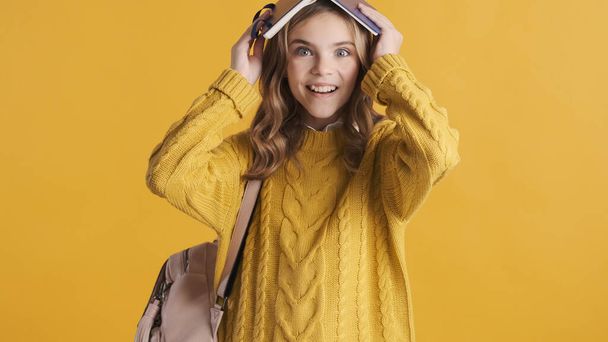 Fröhliches blondes Teenager-Studentenmädchen mit Buch auf dem Kopf, das fröhlich isoliert auf gelbem Hintergrund aussieht. Lustige Schülerin - Foto, Bild