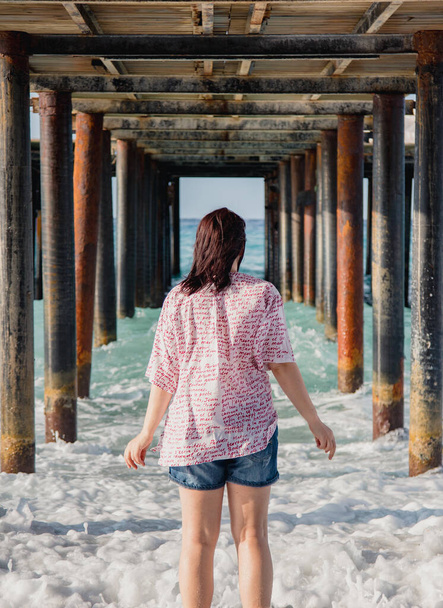 Turkije, Middellandse Zee. Jonge vrouw draaide zich weg van de camera, weglopen van de golven onder een houten pier. - Foto, afbeelding