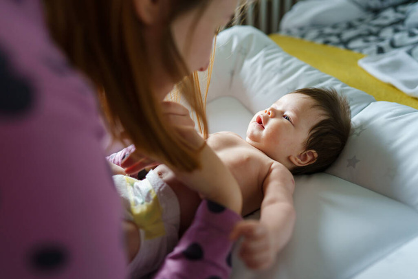 Over the shoulder view of baby half naked two months old boy or girl lying happy on the bed while her mother is near taking care growing up parenthood and motherhood love and bonding concept - Foto, imagen