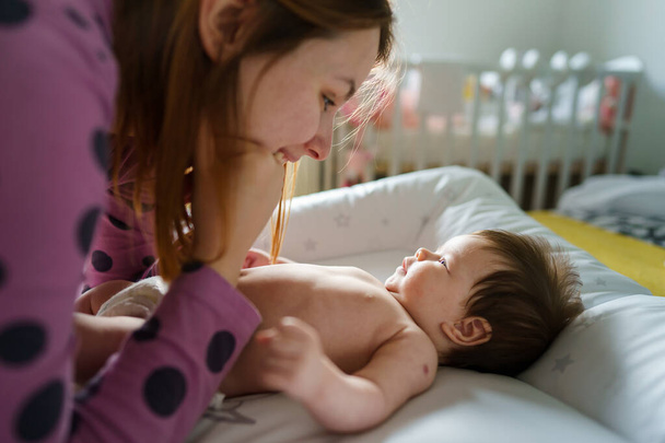 Annesi ona bakarken ve ebeveynlik, annelik, sevgi ve bağlılık kavramı ile ilgilenirken iki aylık bebeğin yan görünüşü yatakta mutlu bir şekilde yatması. - Fotoğraf, Görsel