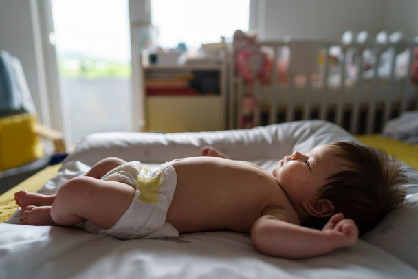 Vista lateral de longitud completa de pequeño bebé caucásico medio desnudo con pañales mientras está acostado en la cama en la habitación en casa en el día - Foto, imagen