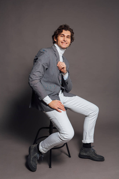 Fashion portrait of a tall handsome man dressed in white turtleneck, white jeans and grey jacket sitting on chair and posing on the grey background - 写真・画像