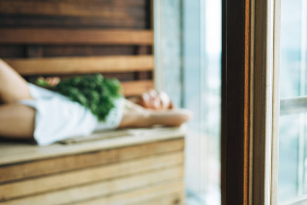 Sumea kuva Nainen valkoisessa pyyhkeessä makaa puisella hyllyllä suomalaisessa saunassa vihreällä tuoreella luudalla, hemmottele itseäsi - Valokuva, kuva