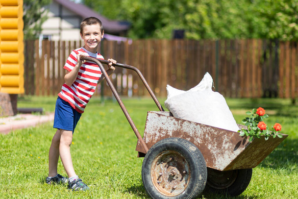 かわいい幼児の男の子は庭の屋外の裏庭で大きな古い車椅子で遊んでいます。若い笑顔の男の子は庭の周りに車椅子を押して. - 写真・画像