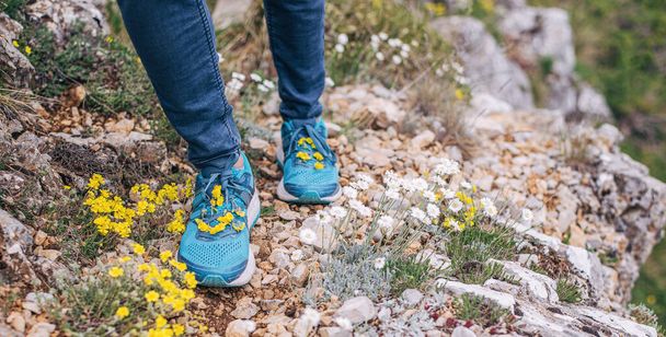 黄色の山の花で飾られた青いスニーカーで岩のハイキングコースの足のクローズアップ。自然と調和して.  - 写真・画像