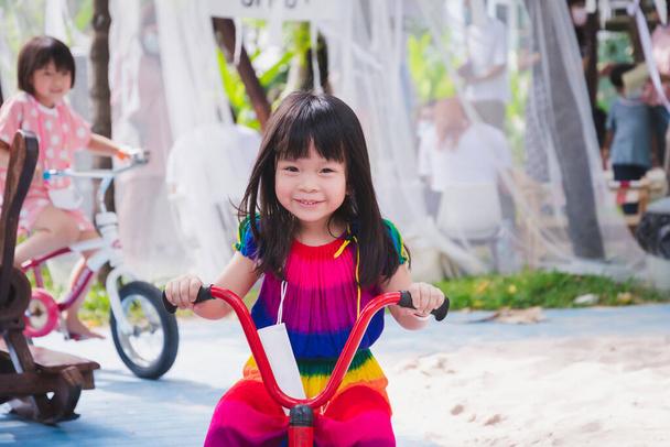 Schattig Aziatisch meisje op een driewieler. Gelukkig kind draagt kleurrijke jurken. Hij glimlacht lief. Kinderen oefenen op de speelplaats. Dame van 4-5 jaar oud. - Foto, afbeelding
