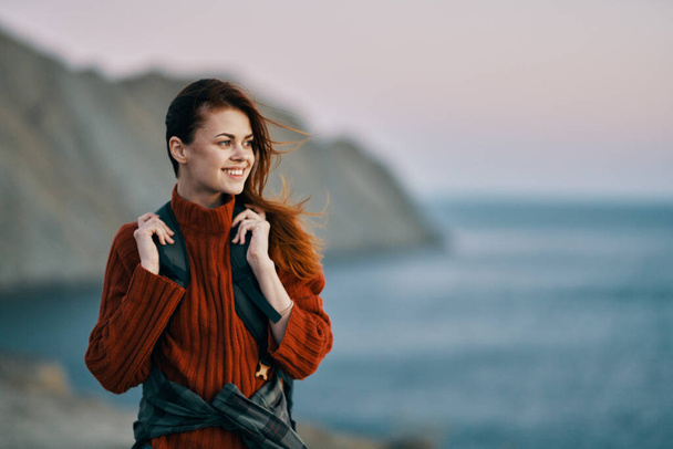 mooie vrouw in een trui houdt een rugzak op haar rug en spreekt in de achtergrond oceaan frisse lucht - Foto, afbeelding