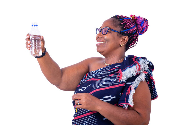 Beyaz arka planda duran geleneksel elbiseli güzel bir Afrikalı kadın bir şişe suyu gülümserken gösteriyor.. - Fotoğraf, Görsel