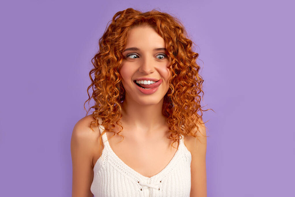 słodkie rude włosy dziewczyna z loki śmieszne wystające z jej język i co twarz jest izolowany na fioletowym tle. - Zdjęcie, obraz