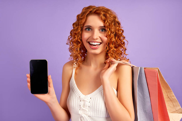 Χαριτωμένο κορίτσι με κόκκινα μαλλιά με μπούκλες και τσάντες ψώνια δείχνει λευκή οθόνη του smartphone που απομονώνονται σε μωβ φόντο. Online αγορές. Πώληση. Σχολιασμός έννοια. - Φωτογραφία, εικόνα