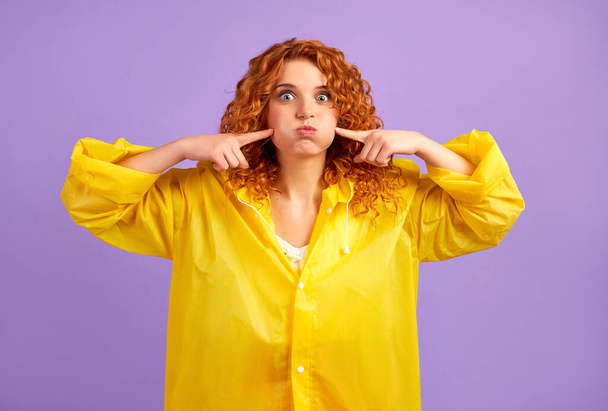 Redhead woman in yellow raincoat having fun isolated on purple background. - Photo, Image