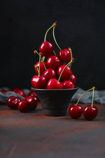 Ciliegie rosse fresche e dolci in una ciotola di terracotta nera su fondo scuro con gocce d'acqua. frutta di stagione, prelibatezza - Foto, immagini