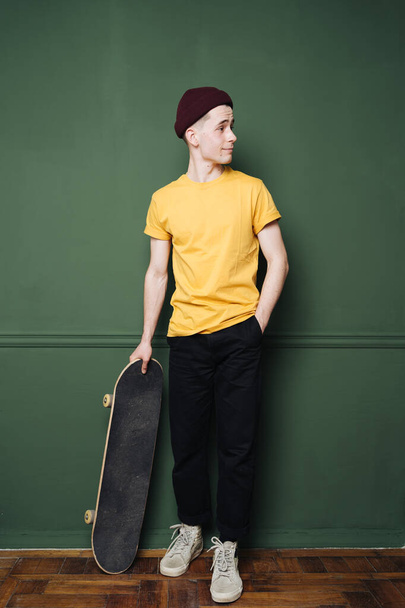 Caucasian man in yellow t shirt posing with skate over dark green background wall - Фото, изображение