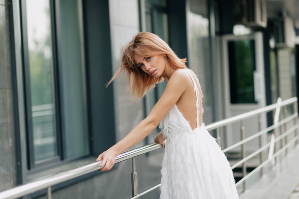 Модная красивая женщина с короткими светлыми волосами одета в белое платье позируя перед камерой в городе. Высокое качество фото - Фото, изображение