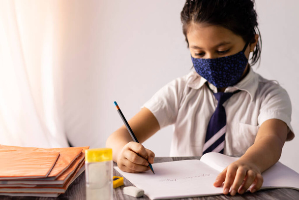 Focus selettivo a portata di mano di un bambino indiano che scrive con la matita sul taccuino indossando uniforme scolastica e maschera nasale - Foto, immagini