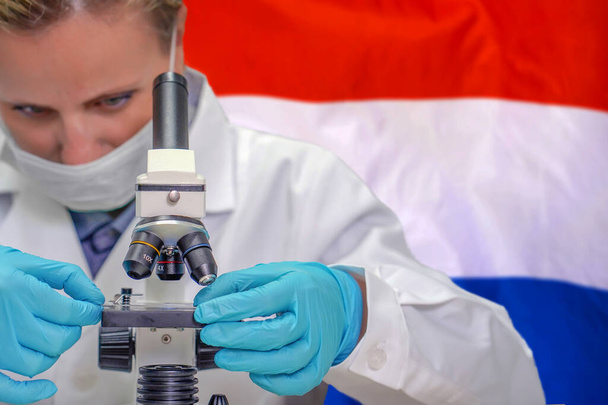 Biochimiste féminine regardant à travers un microscope sur fond de drapeau néerlandais. Technologie médicale et recherche pharmaceutique et développement de concepts scientifiques en néerlandais - Photo, image