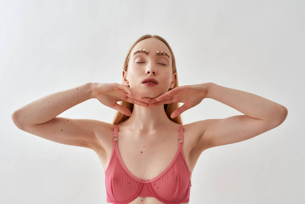 Portrait of sensual young caucasian woman with flowers in her eyebrows wearing pink transparent underwear standing, posing with eyes closed isolated over light gray background - Foto, Imagem