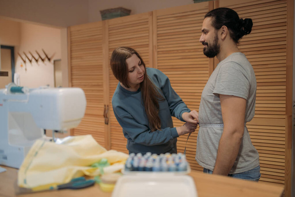 Eine junge Frau nimmt Maße von einem Mann an, um Kleidung zu nähen. Schneiderin bei der Arbeit mit einem Kunden in einer Nähmaschinenwerkstatt - Foto, Bild
