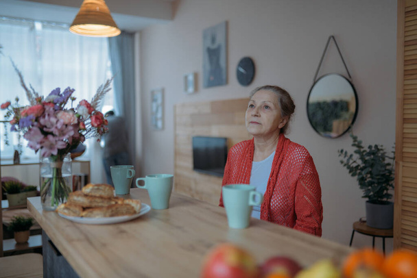 Портрет літньої жінки з квітами за столом. Красива жінка у віці усвідомленого сидіння на кухні вдома у відпустці
 - Фото, зображення