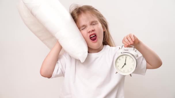 小さなあくびをする少女が白い背景に枕と目覚まし時計を抱いている。幼児期に学校や幼稚園に通っていました。快適な寝具. - 映像、動画
