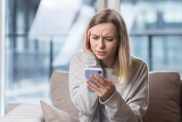 Young woman sitting at home on the couch with a severe headache, trying to call a doctor with a phone, and find a solution to the problem in an online consultation - Foto, imagen