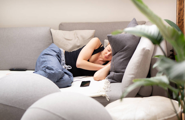 Mujer joven cansada tomando una siesta en casa acostada en un cómodo sofá con grandes cojines en la sala de estar con un teléfono móvil acostado junto a ella. - Foto, imagen