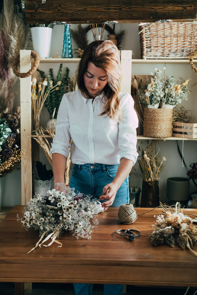 Γυναίκα Ανθοπωλείο κάνει παντοτινή μπουκέτο αποξηραμένα λουλούδια στο ξύλινο τραπέζι στο εργαστήριό της Νεαρή γυναίκα επιχειρηματίας τρέχει τη μικρή επιχείρησή της με ξηρά φυτά και λουλούδια στο ρουστίκ κατάστημα. - Φωτογραφία, εικόνα