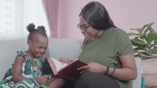 Junge afrikanische Mutter und Tochter beim gemeinsamen Geschichtenerzählen auf dem Sofa zu Hause, Mutter beim Vorlesen mit kleinem Kind auf der Couch im Wohnzimmer mit gemütlichem Familien- und Bildungskonzept. - Filmmaterial, Video
