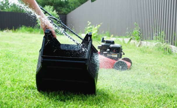 芝刈り機の芝キャッチャーを水で洗う。緑の芝生の背景に水のスプラッシュ。バックグラウンドでガス駆動の自己推進芝刈り機. - 写真・画像