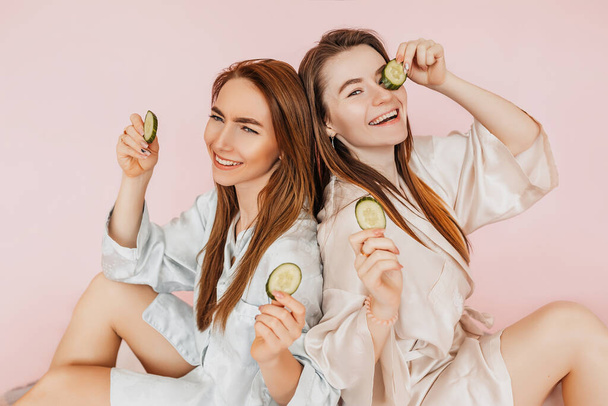 Two girls make homemade face beauty masks. Cucumbers for the freshness of the skin around the eyes. Women take care of youthful skin. Girlfriends laughing and having fun in spa on pink background - Фото, зображення