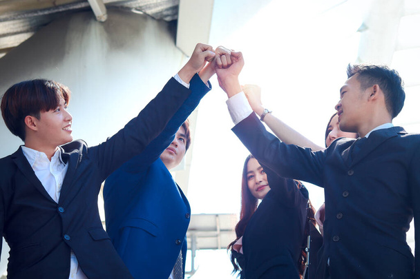 5人の若いアジア人ビジネスマンのグループが手をつなぎ、ダウンタウンのオフィスで手を上げて、団結力のあるチームワークのコンセプトのコミットメントとして手をつないでいます. - 写真・画像