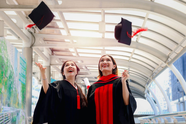 Dwie szczęśliwie uśmiechnięte absolwentki, młode, piękne Azjatki świętujące udaną edukację, świętujące rzucając kwadratową czapkę akademicką w dzień ukończenia studiów (dzień rozpoczęcia), kiedy ukończyły studia na uniwersytecie lub w college 'u. - Zdjęcie, obraz