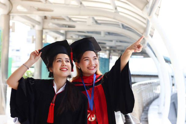 Zwei glücklich lächelnde Hochschulabsolventen, junge schöne asiatische Frauen, die in dieselbe Richtung blicken und etwas zeigen, so stolz auf ihren Starttag, Menschen, die am Abschlusstag eine erfolgreiche Ausbildung feiern, wenn sie ihr Studium an der Universität oder der Universität abgeschlossen haben. - Foto, Bild