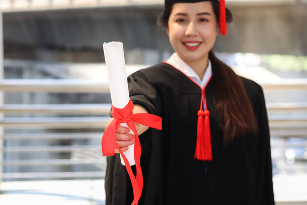 Счастливая улыбающаяся выпускница, молодая красивая азиатка, согревающая квадратную шляпу и дающая сертификат на камеру, отмечающая успешное образование в день начала обучения, когда закончит учебу в университете или колледже. - Фото, изображение