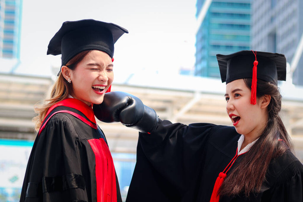 Two happy graduated women students warming square academic hat cap with red boxing gloves or mitt punch, the metaphors about life: life is a fight for territory after celebrating successful education on commencement graduation day. - Photo, Image