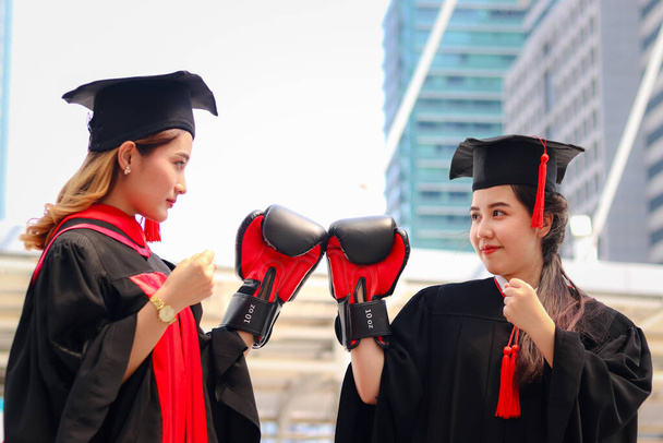 Две счастливые выпускницы студентки согревают квадратные академические шляпы красными боксерскими перчатками или перчатками, метафоры о жизни: жизнь - это борьба за территорию после празднования успешного образования в день выпуска. - Фото, изображение