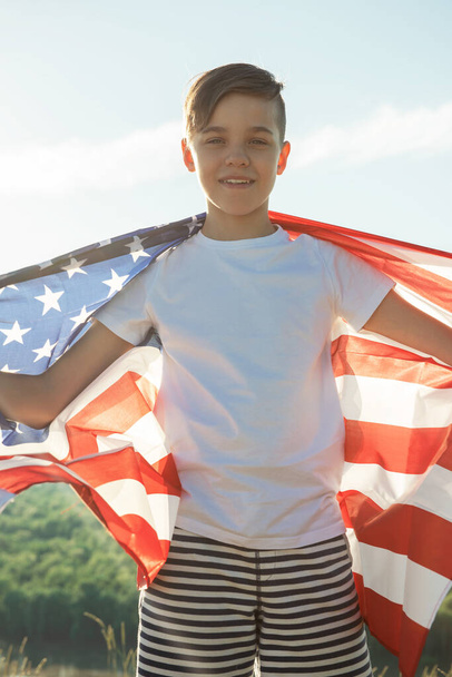 Ξανθό αγόρι κυματίζει εθνική σημαία των ΗΠΑ σε εξωτερικούς χώρους πάνω από τον γαλάζιο ουρανό στην όχθη του ποταμού - Φωτογραφία, εικόνα
