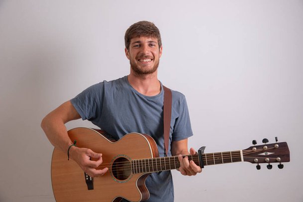 Porträt eines jungen Musikers, der Gitarre spielt und lächelt. Studiofoto eines Künstlers isoliert auf weißem Hintergrund. Junger lächelnder Mann. - Foto, Bild