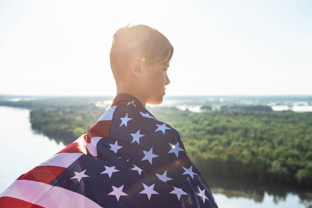 Niño rubio ondeando bandera nacional de EE.UU. al aire libre sobre el cielo azul en la orilla del río - Foto, Imagen