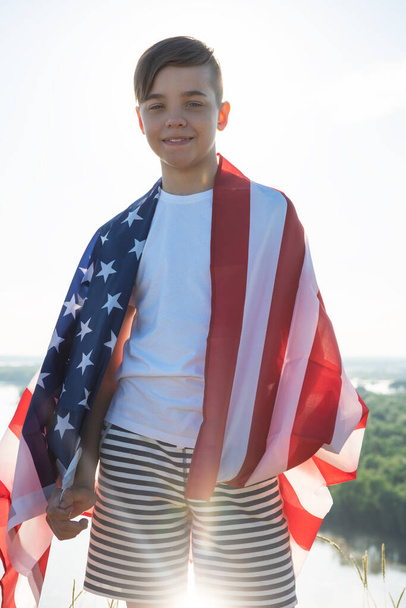 Ragazzo biondo sventola bandiera nazionale USA all'aperto sopra il cielo blu sulla riva del fiume - Foto, immagini