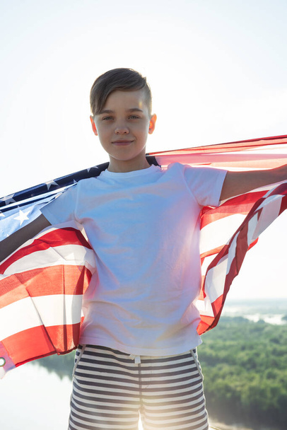 Ξανθό αγόρι κυματίζει εθνική σημαία των ΗΠΑ σε εξωτερικούς χώρους πάνω από τον γαλάζιο ουρανό στην όχθη του ποταμού - Φωτογραφία, εικόνα