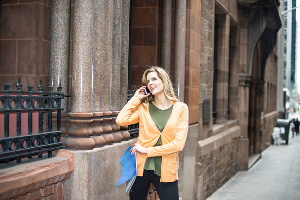 Sujetando una revista y de pie afuera en una calle una joven mujer bonita está haciendo una llamada telefónica  - Foto, imagen