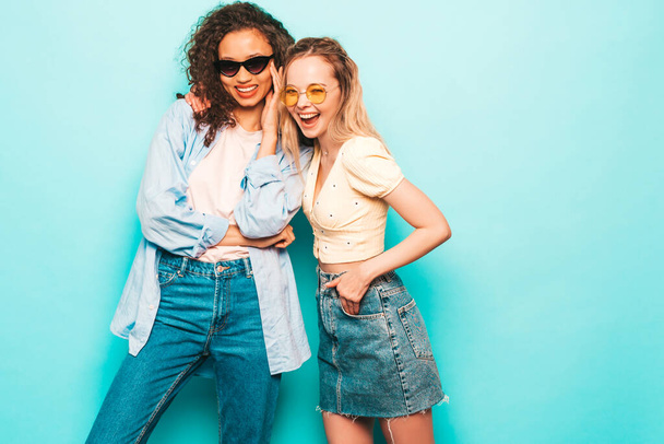 Two young beautiful smiling international hipster female in trendy summer clothes. Sexy carefree women posing near blue wall in studio. Positive models having fun. Concept of friendship - Valokuva, kuva