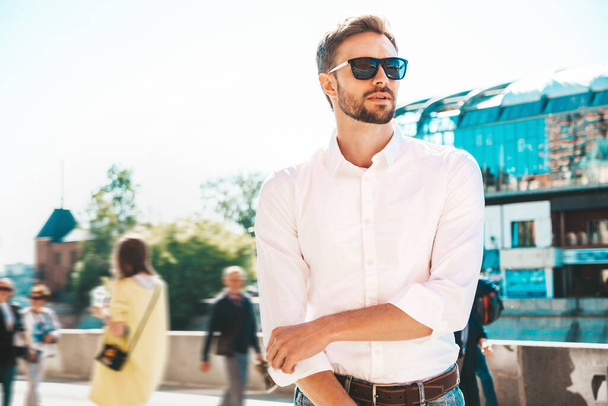 Portrait of handsome confident stylish hipster lambersexual model.Modern man dressed in white shirt. Fashion male posing on the street background in sunglasses. Walking outdoors at sunset  - Φωτογραφία, εικόνα