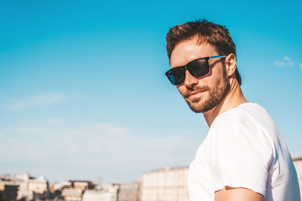 Closeup portrait of handsome smiling  hipster lambersexual model.Stylish man dressed in white T-shirt. Fashion male posing behind blue sky on the street background in sunglasses - Fotó, kép