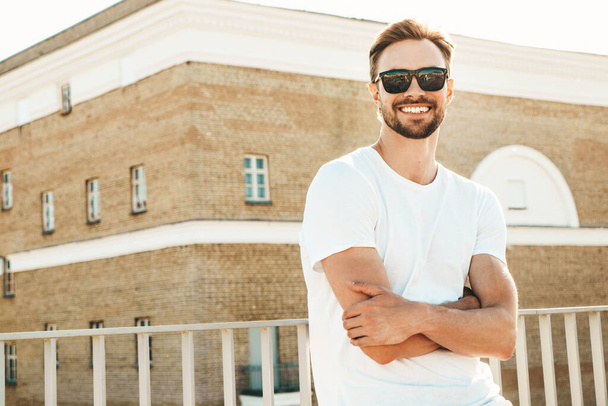 Portrait of handsome smiling  hipster lambersexual model.Stylish man dressed in white T-shirt and jeans. Fashion male posing on the street background near fence at the beach in sunglasses - Fotografie, Obrázek