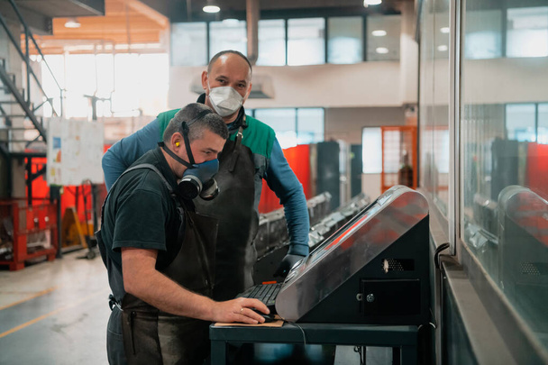 Двое рабочих в маске из-за пандемии коронавируса программируют современную машину с ЧПУ. Высокое качество фото - Фото, изображение